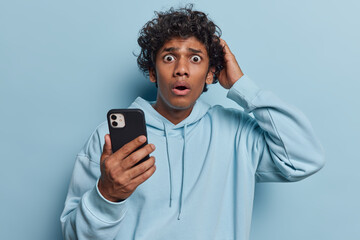 Indoor shot of shocked curly Hindu man keeps hand on head holds mobile phone and chats online...