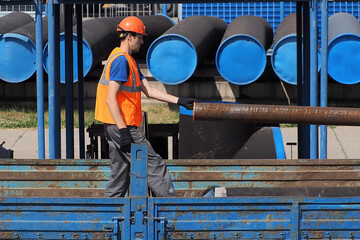 Slinger in helmet and vest unloads metal pipes outside on summer day. Loading metal pipes for...