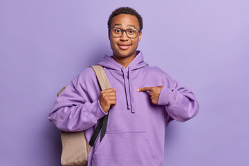 Studio shot of dark skinned African man with curly hair points at himself asks who me dressed in casual sweatshirt poses with backpack surprised being chosen isolated over purple background.
