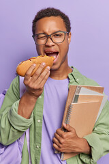 Vertical shot of dark skinned man with short curly hair eats hot dog and carries notepads has lunch...