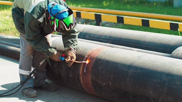 A welder in a mask and overalls cuts a metal pipe with an oxygen torch outdoors on a summer day. Scrap metal recycling.