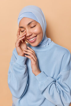 Vertical image of pleased Islamic woman keeps eyes closed touches face gently smiles broadly wears traditional hijab and blue long sleeved jumper isolated over brown background recalls something