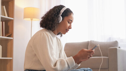 Mobile message. Happy woman. Network connection. Smiling female in headphones chatting smartphone sitting cozy home living room.