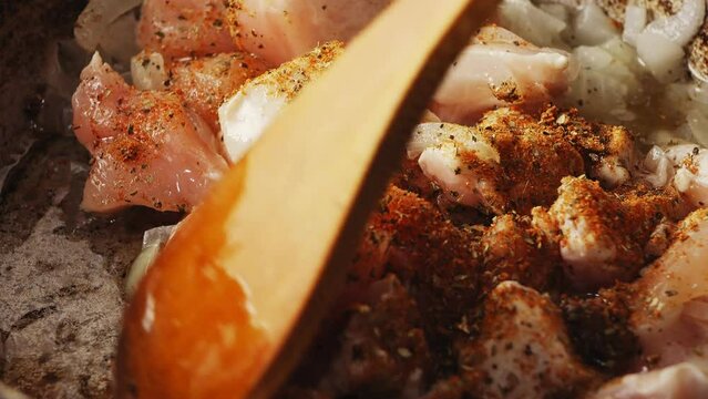 Pieces of chicken fried in a cast-iron skillet in oil with onions and spices are mixed with a spatula. Close-up