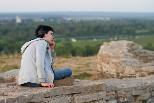 A teenage boy with headphones listening to music on the ruins of an ancient fortress. In the background is a blurry image of the cityscape.