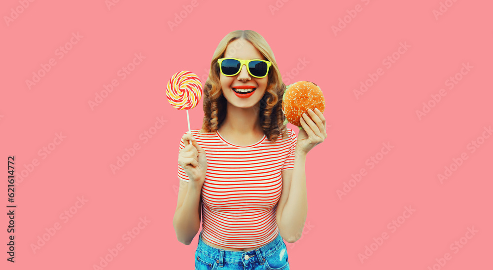 Wall mural Portrait of cheerful young woman with burger fast food and colorful lollipop on pink background