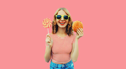 Portrait of cheerful young woman with burger fast food and colorful lollipop on pink background
