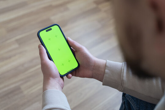 Man at home lying on a couch using smartphone with green mock-up screen, doing swiping, scrolling gestures. Guy using mobile phone, internet social networks browsing. Social media.