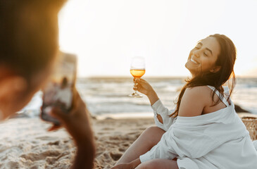 Woman taking a photo of her happy girlfriend at the beach, having picnic and drinking wine at sunset on coasline