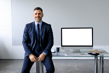 Fototapeta premium Happy middle aged businessman in formal suit sitting at table with blank computer monitor, offering mockup for design