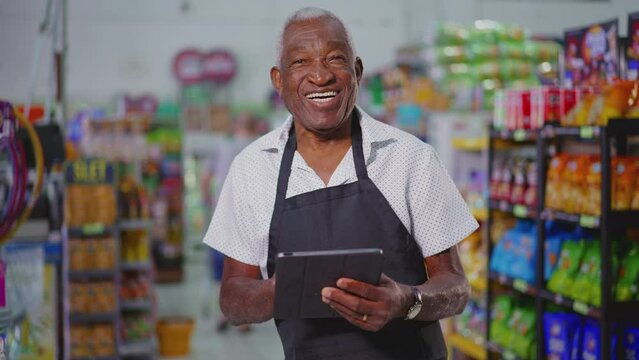 Joyful Happy Brazilian senior employee of supermarket wearing apron and holding tablet device, portrait of a black older male staff depicting job occupation at retail store