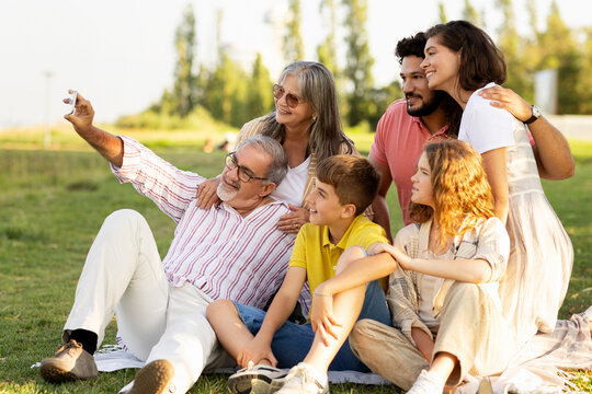 Smiling european old, millennial and teenager family have fun on picnic, take selfie on smartphone in park