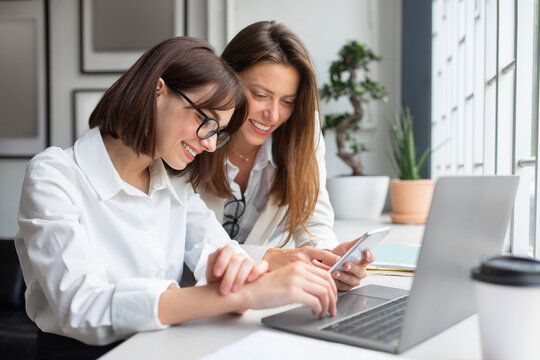 Happy female coworkers looking at smartphone, discussing new e-business application while working in coworking space