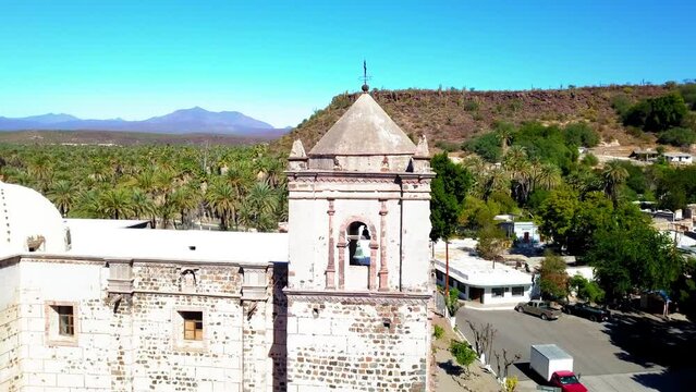 Aerial Panning Shot Of Mission San Ignacio Kadakaaman By Road And Trees, Drone Flying Over Catholic Church Against Blue Sky - Baja California, Mexico
