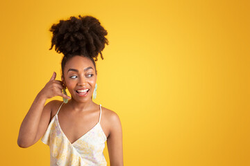 Fototapeta premium Cheerful millennial african american lady in dress making call me gesture, flirting, isolated on yellow background