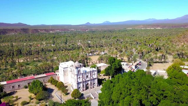 Aerial Shot Of Mission San Ignacio Kadakaaman By Trees, Drone Descending Forward Over Catholic Church On Sunny Day - Baja California, Mexico