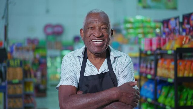 Portrait of a joyful African American senior employee of supermarket wearing apron and smiling at camera inside grocery store aisle and arms crossed