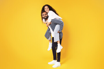 Arabic Man Giving Piggyback Ride To Wife Fooling, Yellow Background