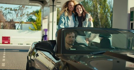 guy and two girls arrived in a gray convertible to a gas station. The guy gets out of the driver's seat and fills up the car