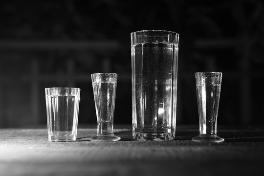 Black and white still picture of collection old vintage glasses consist from big glass for water, thin shot glasses and one standard shot glass. Picture is taken in the dark, outside on the terrace.