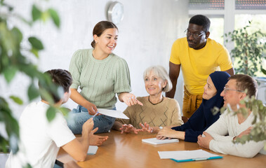 Interested friendly young girl, student of adult school, taking part in discussion of study topics with international mixed age group of coursemates sitting at table..