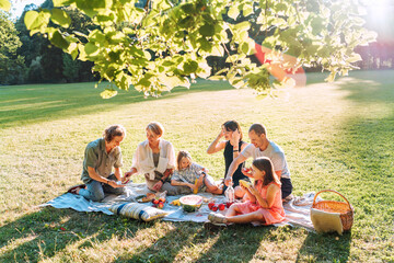 Big family under Linden tree on the picnic blanket on the in city park green grass. They are eating boiled corn, apples, peaches, pastries and watermelon. Family values and outdoor activities concept.