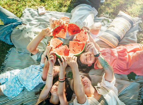 Young daughters with parents family lying on picnic blanket during weekend sunny day, smiling, laughing and rose up red juicy watermelon pieces. Family values, fruits vitamins, outdoor time concept