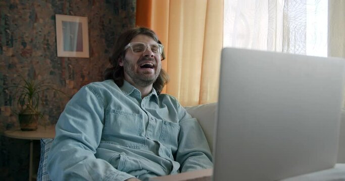 Young man freelancer laughing hard, watching entertaining funny content on his laptop online. Laugh with sarcasm, irony. Man with long hair, wearing glasses at home. Mood change