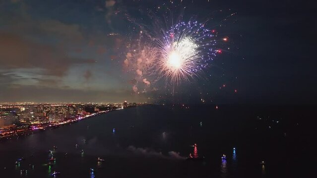 Aerial view of fireworks launching from the barge at night by the beach. City lights on the horizon