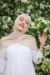 A young Muslim woman in a white dress in front of a flowering tree. Medium plan. Blooming apple tree. Pretty modest Muslim woman. European Muslim. Tenderness. Beauty.