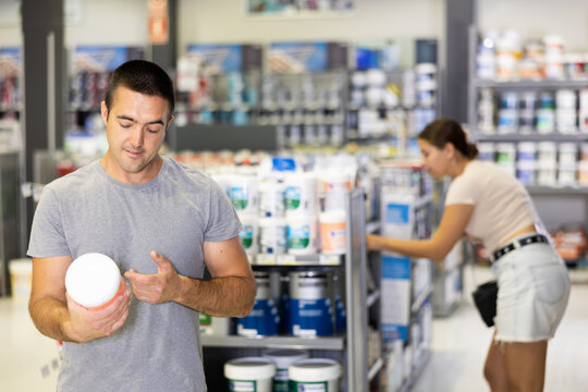 Positive interested young man attentively reading labels on paint cans, choosing high-quality materials for apartment renovation at hardware store