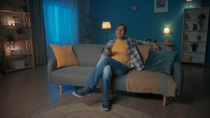 Obraz premium A man sits on the sofa in the evening and watches TV. A man changes channels with a remote control.