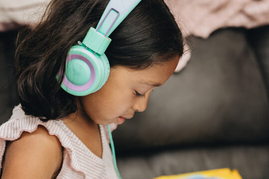 diverse kindergartner girl at home on sofa looking at tablet computer with headphones