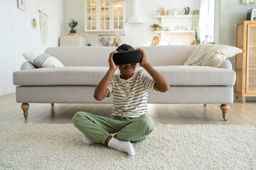 African American boy child sitting on floor in living room wearing virtual reality headset exploring virtual world. Schoolboy kid using VR technology for learning. Augmented reality for children