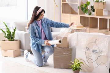 Young woman wrapping cardboard box with stretch film at home on moving day