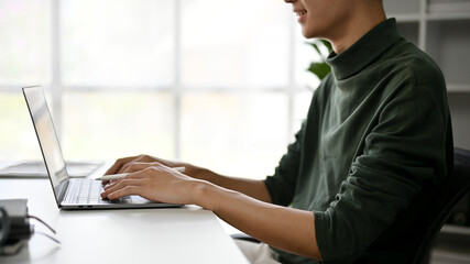 An Asian man typing on keyboard, using a laptop computer at his desk in a modern office.