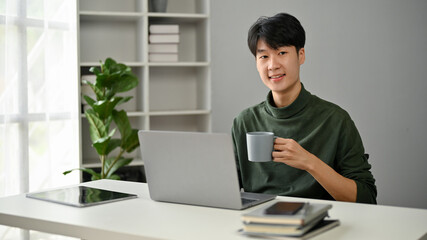 A successful Asian man sipping coffee at his desk in a modern office room.