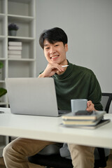A portrait of a handsome Asian man in causal clothes sits at his desk in a modern office alone.