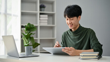 A happy Asian man using his digital tablet to manage his work in his home office.