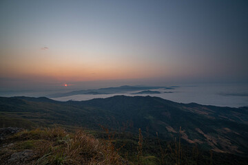 The stunning view from a tourist's standpoint as they go down a hill on a foggy trail with a hill and a background of a golden sky in Phu Langka Forest Park in Phayao, Thailand.