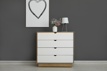 Stylish white chest of drawers with gypsophila flowers, lamp and picture hanging on grey wall in room