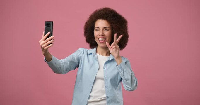Smiling happy arican american woman in jeans shirt making selfie on smartphone over pink studio background