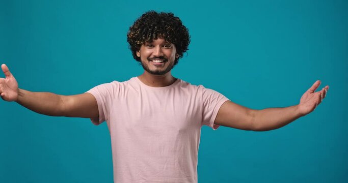 Young charismatic smiling happy cheerful indian bearded man wears pink t-shirt reach out hands to camera say come give me hug standing isolated blue studio background