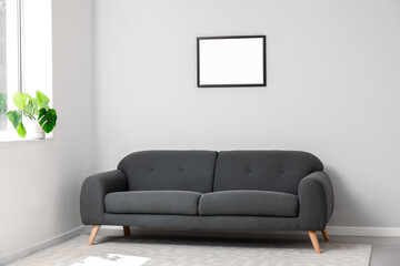 Interior of light living room with cozy grey sofa, houseplant on windowsill and blank frame