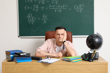 Tired male teacher sitting at table in classroom