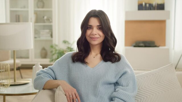 Pleasant calm latin woman feels satisfied posing for portrait view in living room modern apartment, slow motion people. Smiling beautiful hispanic woman looking at camera sitting on sofa alone at home