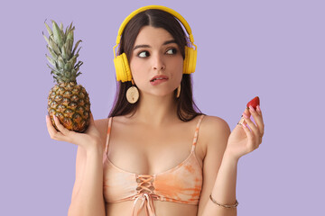 Thoughtful young woman in headphones with pineapple and strawberry on lilac background