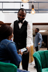 Fototapeta na wymiar Smiling African American man waiter giving pos machine to female hotel guest sitting in lobby. Tourist holding smartphone paying for order while waiting for check-in procedure at lounge area
