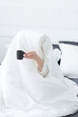 Young woman with cup of coffee sitting in bed under white blanket at home bedroom in the morning. Daily routine, early wakening.
