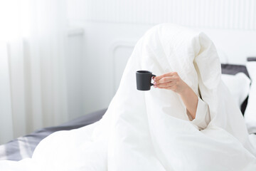 Young woman with cup of coffee sitting in bed under white blanket at home bedroom in the morning. Daily routine, early wakening.